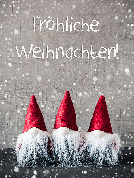 Drei dekorative Weihnachtswichtel mit roten Zipfelmützen und langen weißen Bärten stehen nebeneinander vor einer grauen Wand. Darüber steht in weißer Schrift ‚Fröhliche Weihnachten!‘. Im Vorder- und Hintergrund sind Schneeflocken zu sehen, die ein winterliches Ambiente erzeugen.